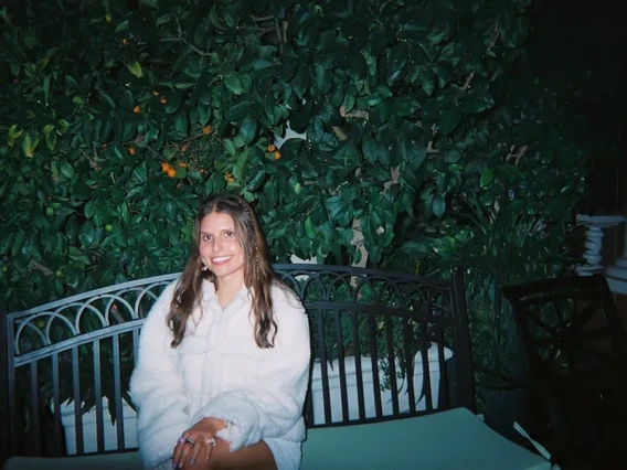 Emerson sitting on a bench in front of a green bush. They are wearing a long-sleeve, buttoned white shirt and smiling at the camera.