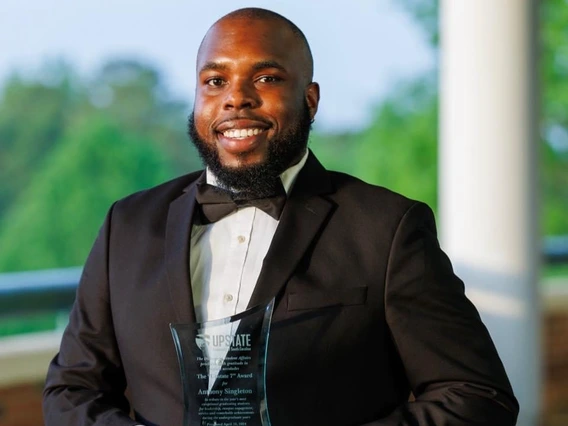 image of smiling Black man wearing a tuxedo and holding a glass plaque. There is greenery behind him and white pillar just over his left shoulder. 