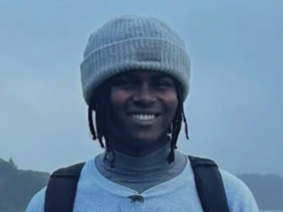 Smiling, young black man with braids wearing a winter cap , a Dallas cowboys tee shirt and a black backpack. There re reflective ray bans hanging from the collar of his tee/.