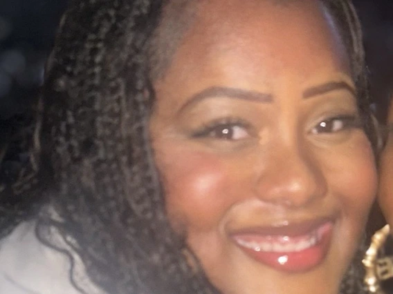Smiling young Black woman with face framing braids and glossy red lipstick. She is wearing a white shirt. The background is dark. 