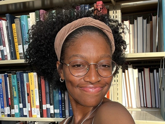 Smiling Black woman (not showing her teeth) with a medium sized afro and a mauve colored headband. She is wearing glasses, looking at the camera and wearing a white tank top. She is posing in front of a library shelf of books.