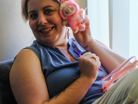 Audrie seated in a window with their legs drawn up, holding a pink and yellow Labubu up by their cheek.