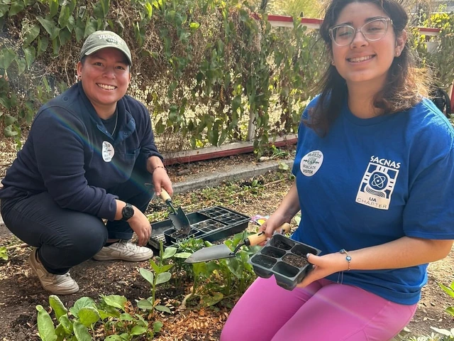 2 students kneeling in a garden holding planters and surrounded by plants