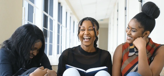 Three black girls sitting on steps laughing. Girl on left has on long sleeve dark shirt and dark jeans and her head is down. SHe has long black hair. Girls in middle has face framed by two braids - she is smiling with her mouth open and has an open book in her lap. She is wearing a dark shirt and white jeans with white sneakers and the girl on the right has her hair in a high bun. SHe has on a multicolored sleeveless mock turtleneck, jeans and black boots.