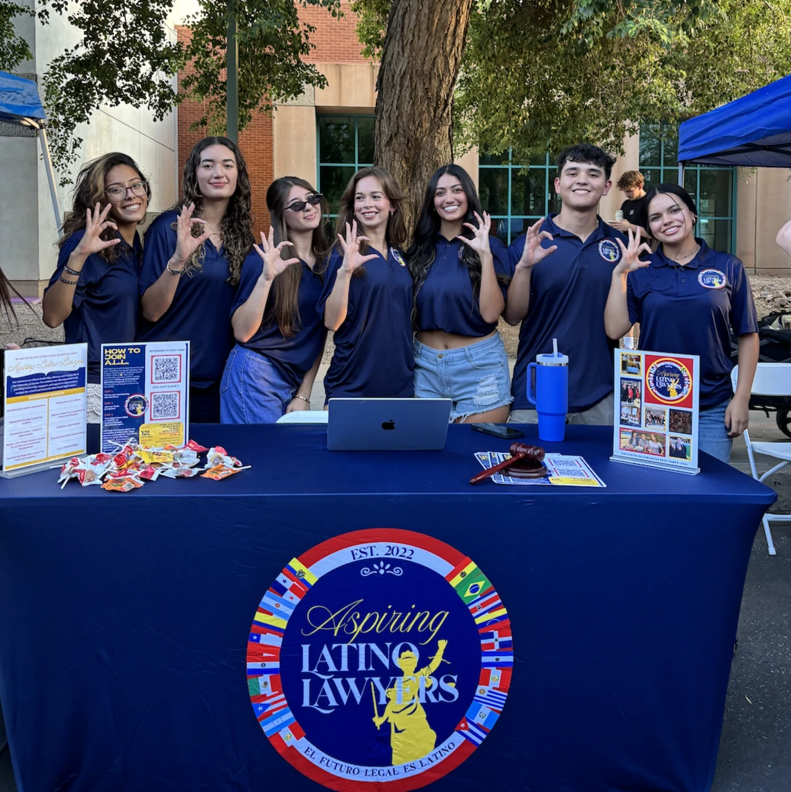 Students standing in front of a table, students are wearing blue polos, they are holding the wildcats sign with their hand