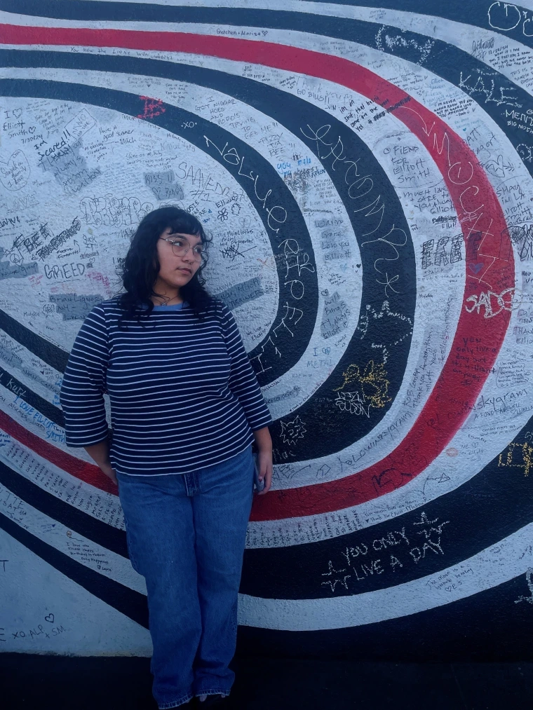 Ashley leaning against a wall with white, navy blue, and red abstract swirls and hand-written messages all over it. She is standing and leaning slightly on her right hip. She is looking off to the left.