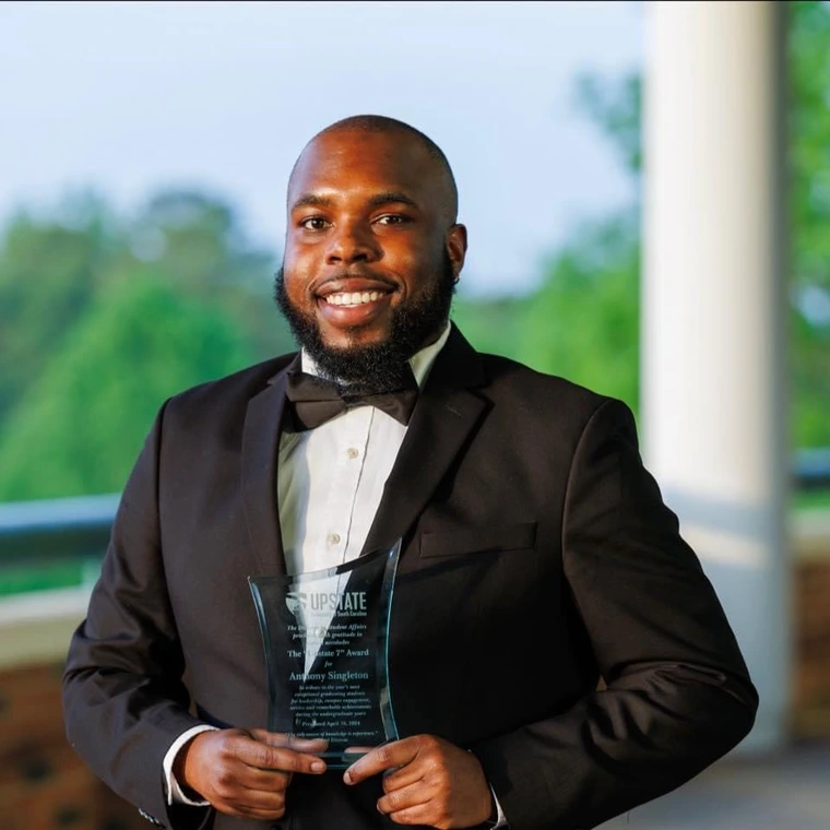 image of smiling Black man wearing a tuxedo and holding a glass plaque. There is greenery behind him and white pillar just over his left shoulder. 