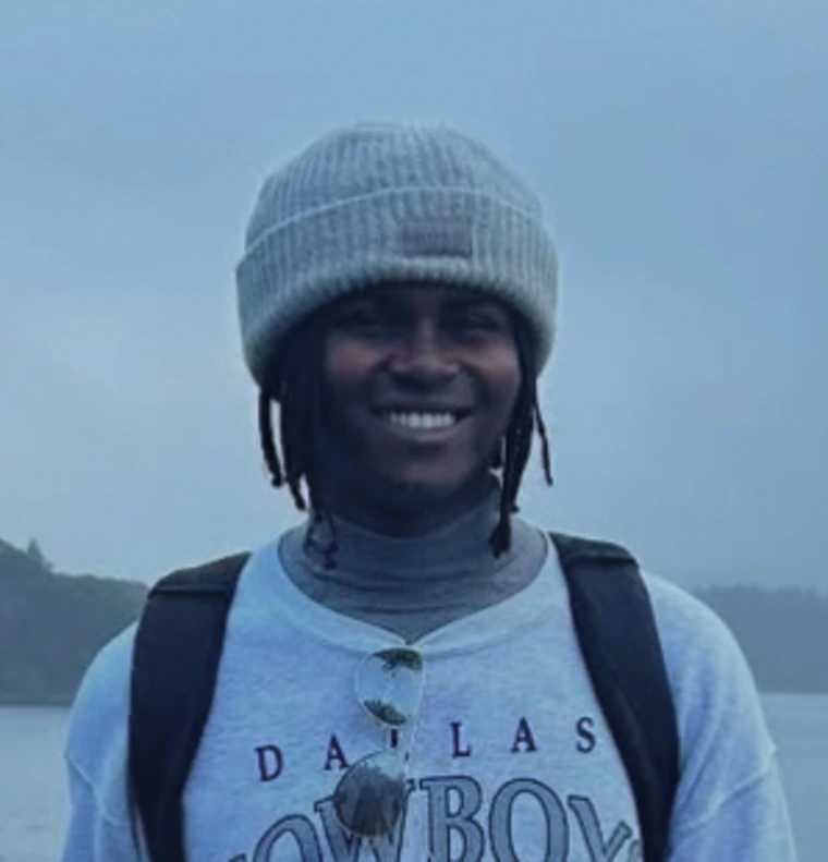 Smiling, young black man with braids wearing a winter cap , a Dallas cowboys tee shirt and a black backpack. There re reflective ray bans hanging from the collar of his tee/.