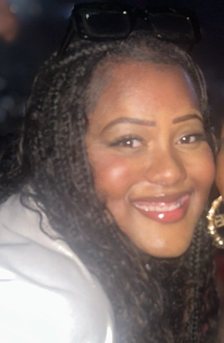 Smiling young Black woman with face framing braids and glossy red lipstick. She is wearing a white shirt. The background is dark. 