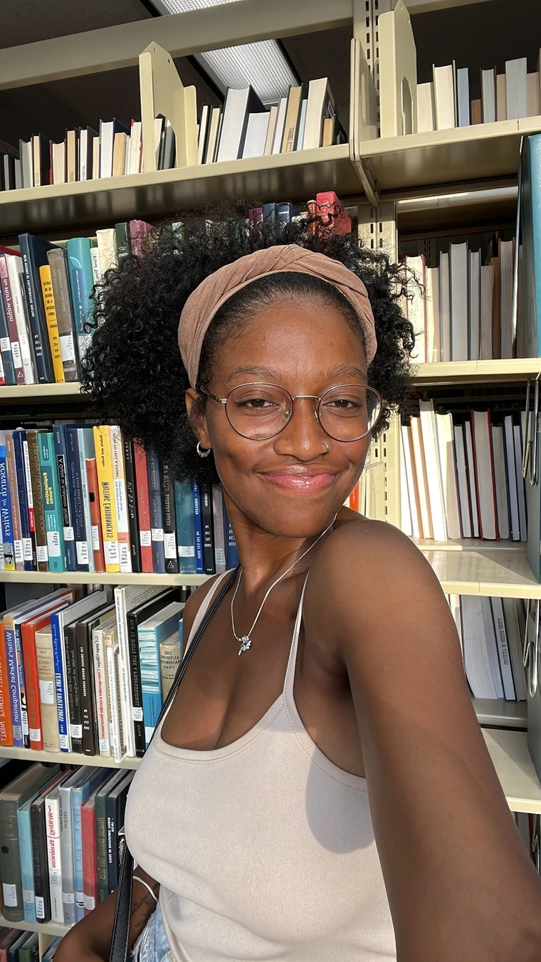 Smiling Black woman (not showing her teeth) with a medium sized afro and a mauve colored headband. She is wearing glasses, looking at the camera and wearing a white tank top. She is posing in front of a library shelf of books.