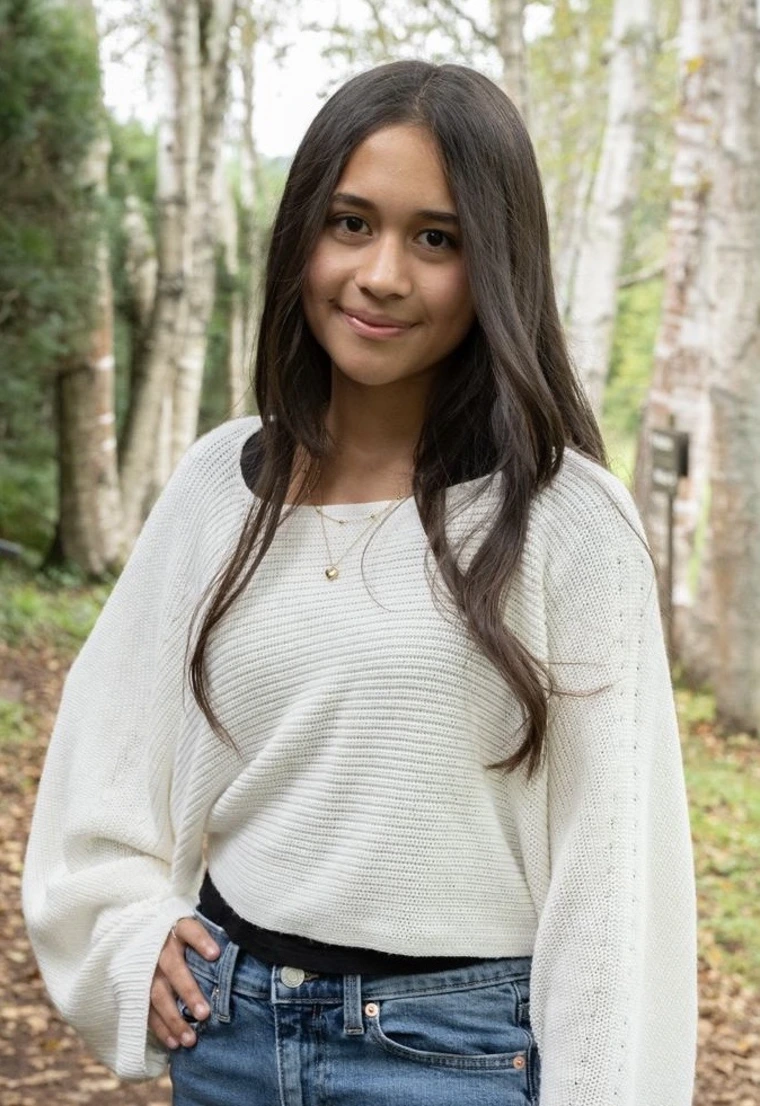 Image of a student with long hair, white sweater, standing in front of trees smiling 