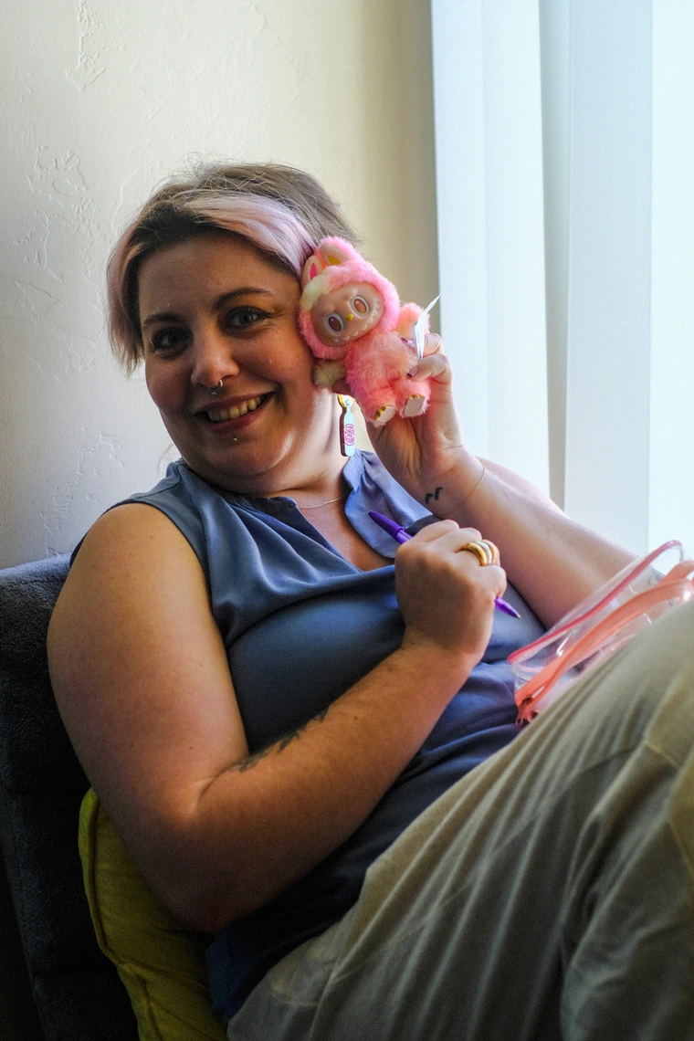 Audrie seated in a window with their legs drawn up, holding a pink and yellow Labubu up by their cheek.