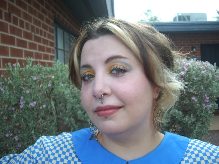 Audrie standing in front of green bushes and a brick wall. They are wearing multi-colored eyeliner and are tilting their head to the side.