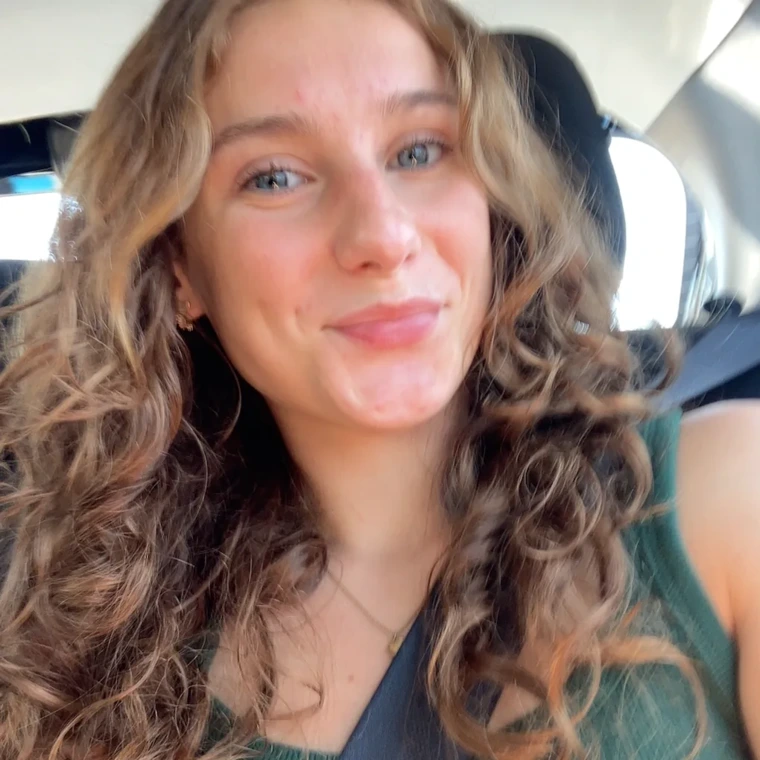 Alex smiling at the camera in a selfie. She is sitting in a car, head slightly tilted with her curly hair pulled over her shoulders.