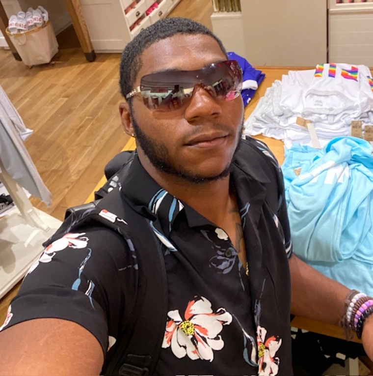 Black male with sunglasses on and a black beach shirt on with flowers. 