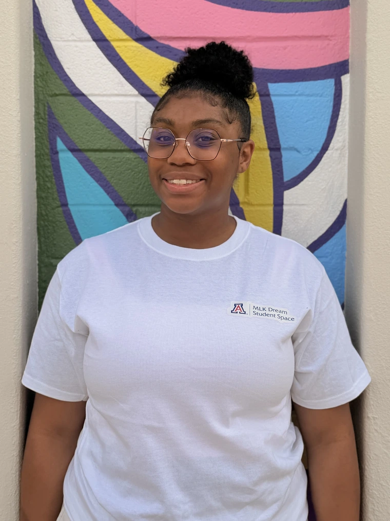 African American female student with hair in a high bun and circular glasses, wearing a white tee shirt that says "MLK Dream Student Space" in the upper left corner