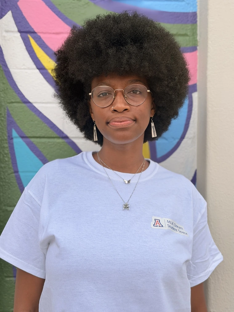 African American female with an afro hairstyle, wearing round glasses. She is smiling but not showing her teeth.  She is also wearing a white shirt that says "MLK Dream Student Space" in the upper left corner.