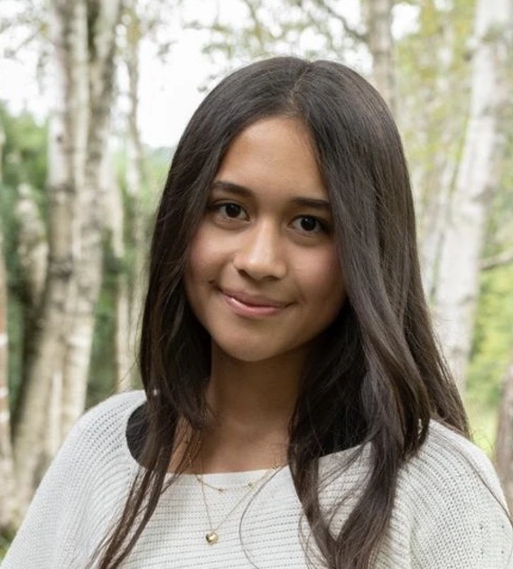Image of a student with long hair, white sweater, standing in front of trees smiling 