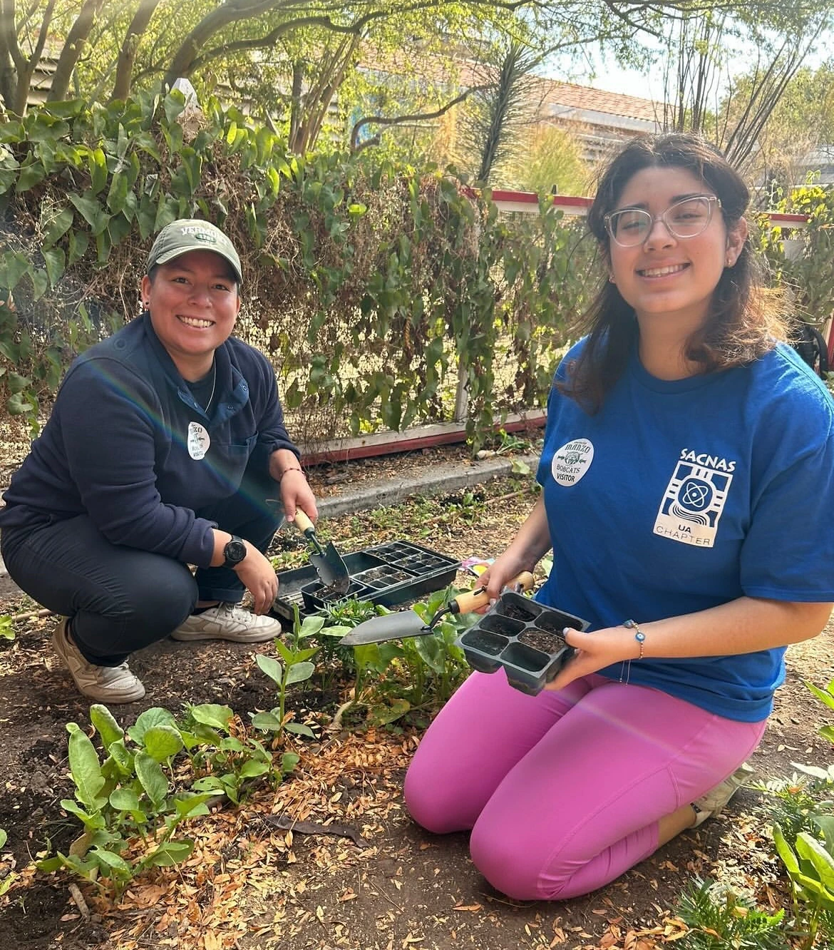 2 students kneeling in a garden holding planters and surrounded by plants
