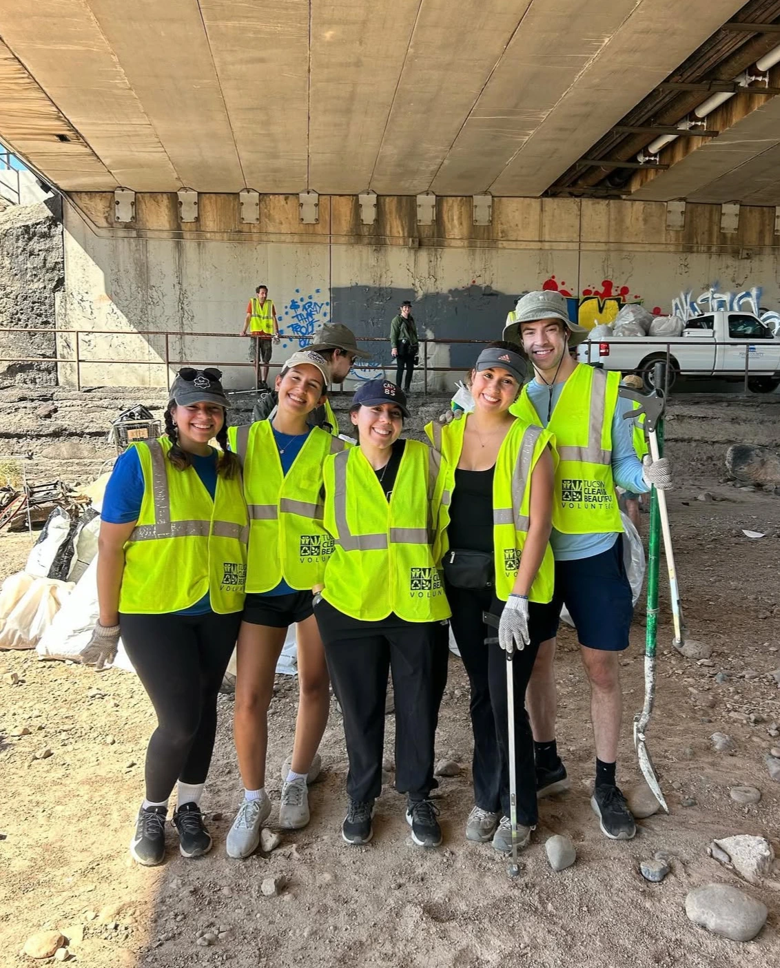 5 students wearing bring yellow vests holding shovels and other tools